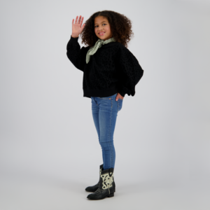 Girl with curly hair in a black leopard-print sweater, light green scarf, blue jeans, and cowboy boots waves hello.