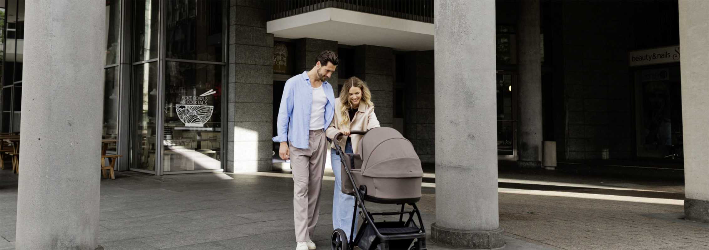 Couple pushes a beige baby stroller down a city sidewalk between tall concrete columns.