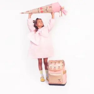 Young girl in a pink dress lifts an oversized pencil above her head, with a pink backpack beside her.