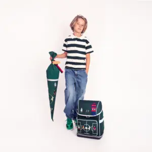 Boy standing in striped polo and jeans holds a rolled umbrella beside a matching backpack and school bag with nautical designs