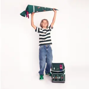 Young boy in a striped shirt and jeans raises a large green wrapped item above his head, with a matching green backpack on the floor nearby.