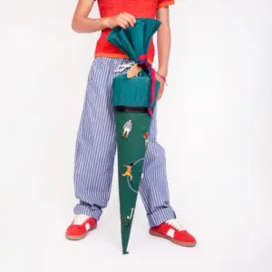 Person in a red shirt and blue striped pants holds a tall green cone-shaped umbrella with sport-themed illustrations and a pink ribbon.