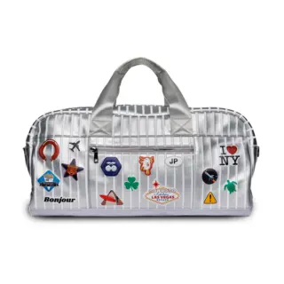 Silver duffel bag with stitched handles and luggage stickers on a white background