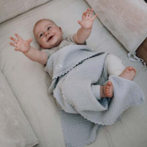 Smiling baby lying on a light sofa with a gray textured blanket draped over the legs, arms raised playfully.