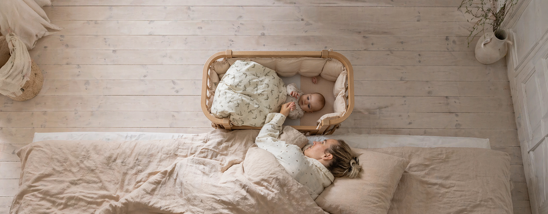Top-down view of a mother in a pink bed reaching toward her baby in an oval wooden bassinet beside the bed.