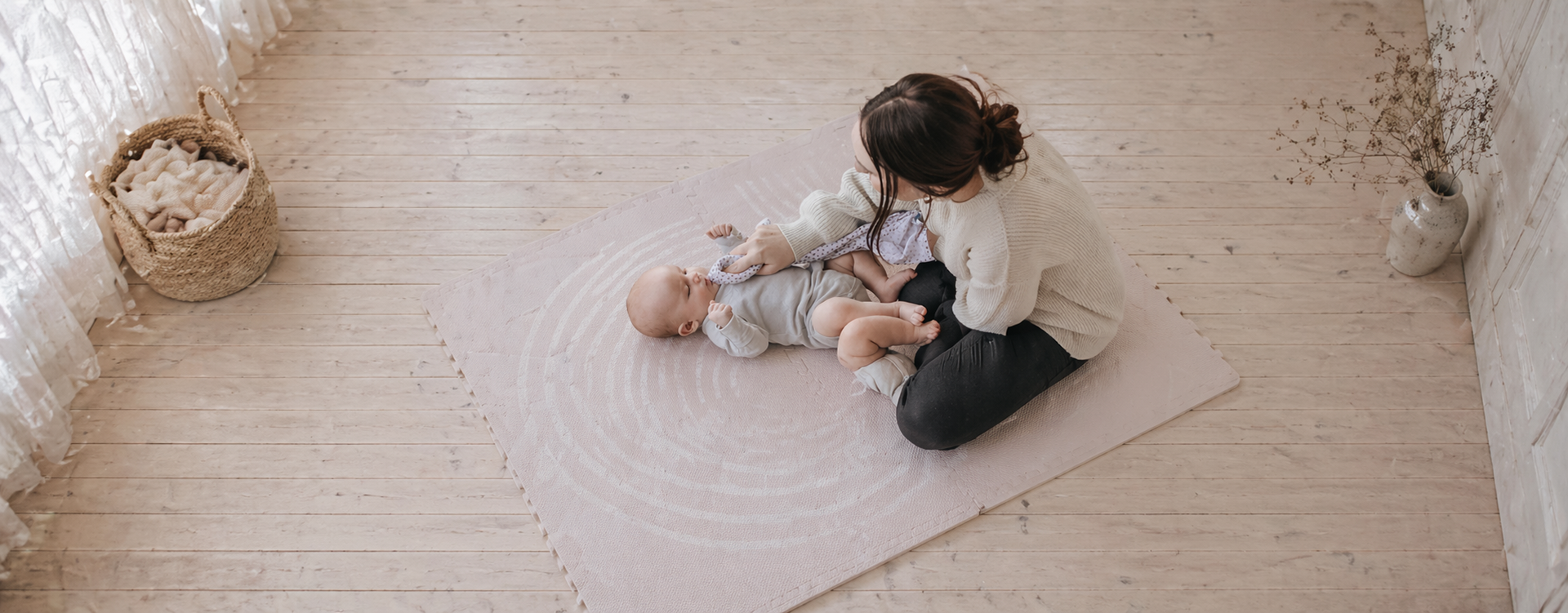 Mother sitting on a pink rug, cradling a newborn; basket of blankets on the left and a vase with branches on the right.