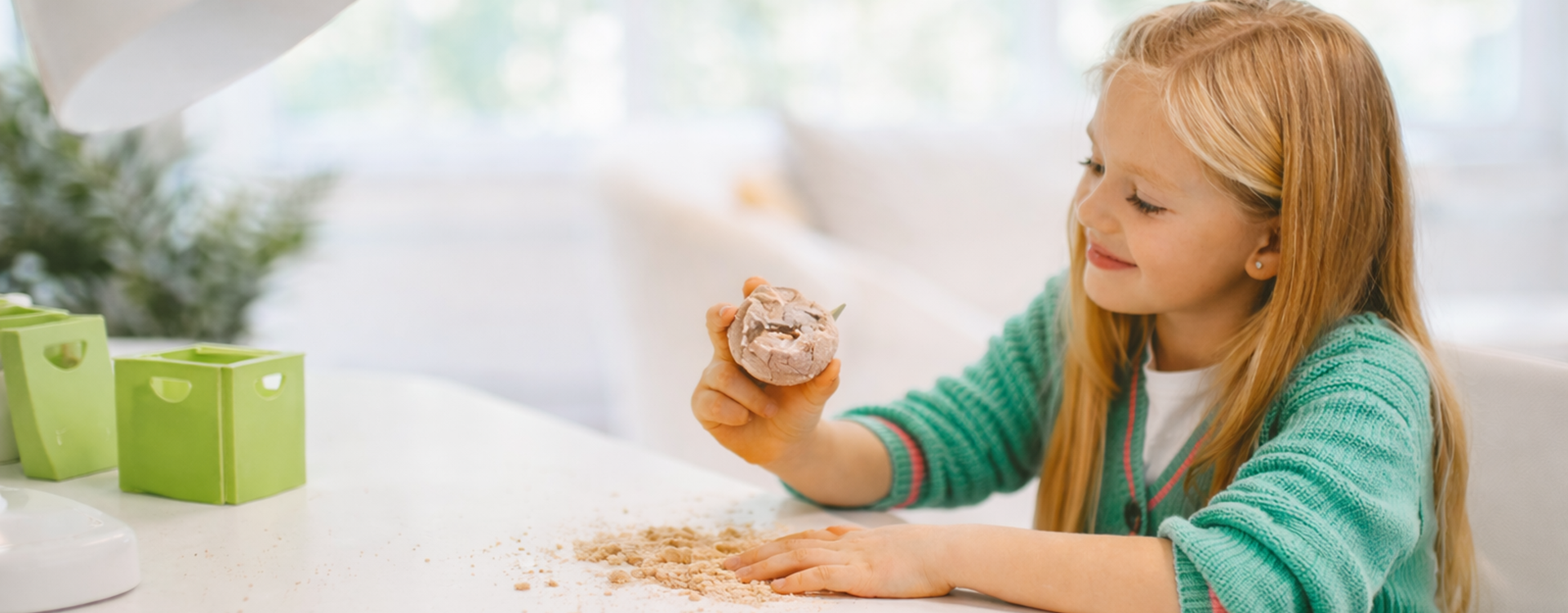 Young girl with blonde hair sits at a white table, shaping a crumbly ball of dough in her hand.