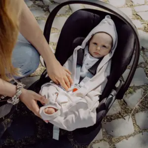 Baby strapped into an infant car seat outdoors while a caregiver adjusts the harness on a stone-paved path.