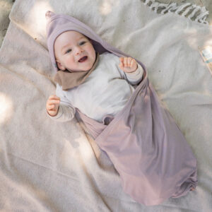 Smiling baby wrapped in a lavender swaddle, lying on a light blanket outdoors.