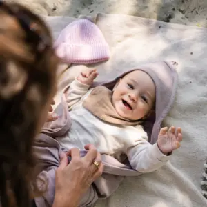 Smiling baby bundled in a light outfit on a beige blanket outdoors, with a lavender hood and pink hat nearby.