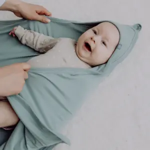 Baby smiling, snug in a mint‑green hooded swaddle being held gentle by an adult.",