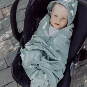 Baby in a light mint-green blanket sits in a stroller on a stone sidewalk, smiling at the camera.