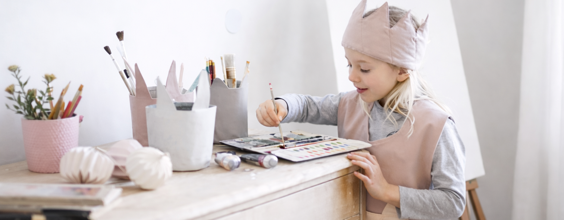 Little girl wearing a pink crown hat paints with a brush at a wooden table filled with art supplies.