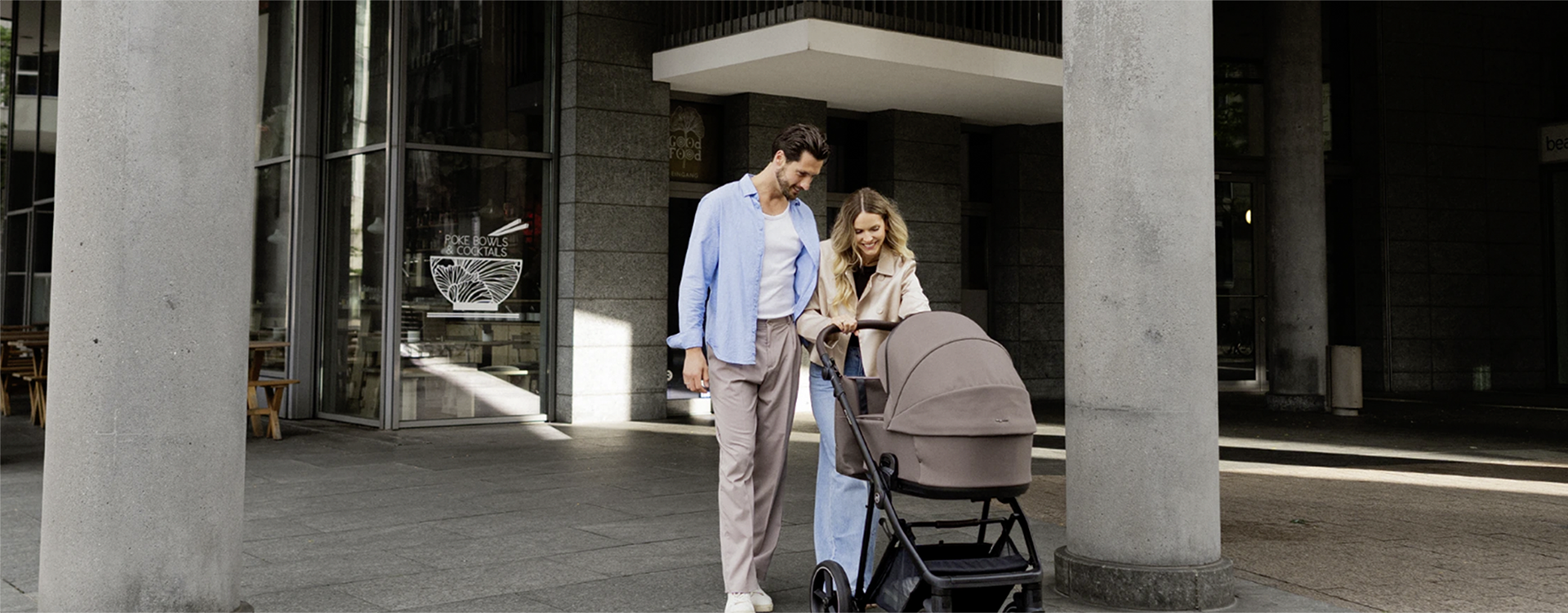 Couple with a baby stroller standing under large concrete columns outside a modern building.