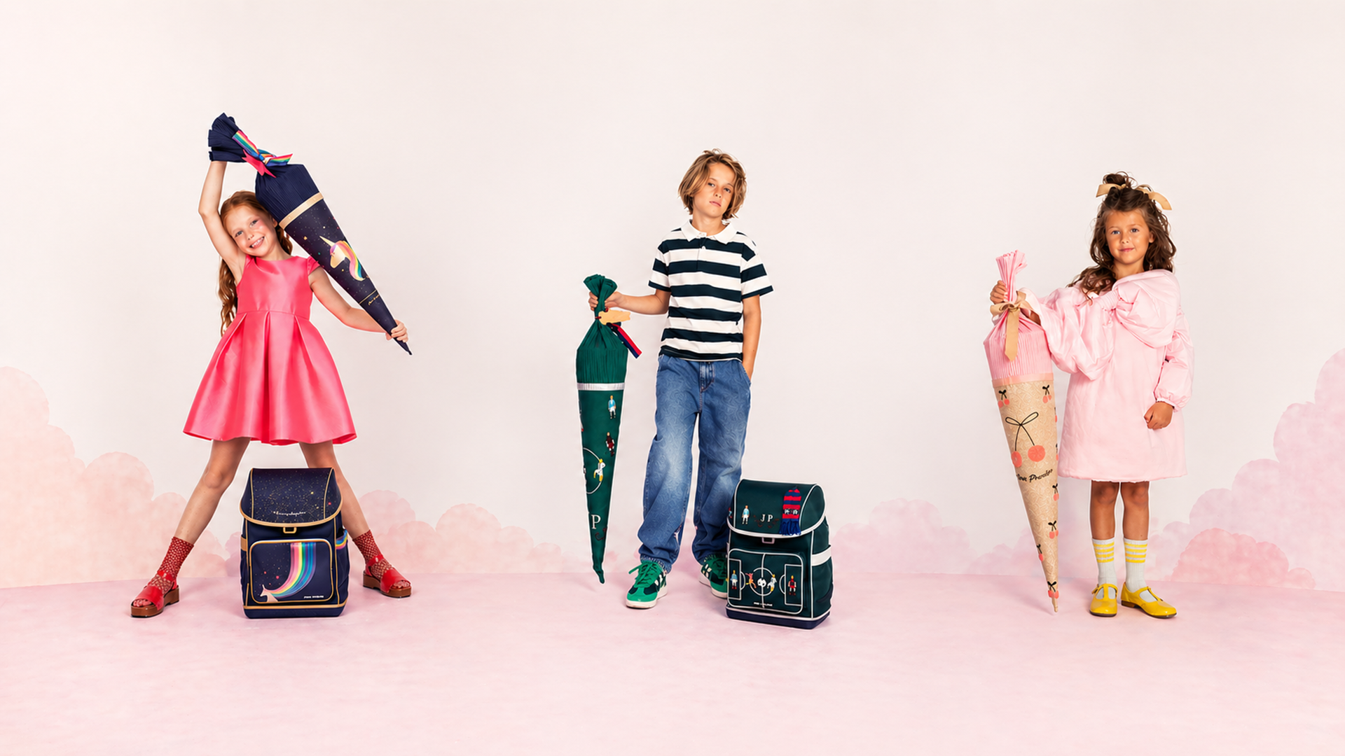 Three kids pose with oversized school supplies and backpacks in a pastel pink scene with clouds in the background.