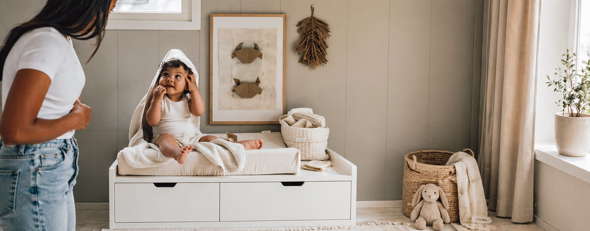 Mother stands beside a white changing table as her baby sits wrapped in a towel on the table in a cozy, neutral-toned nursery.