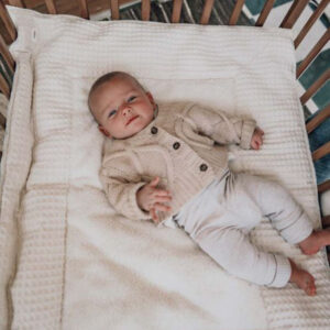 Baby lying on a white blanket in a crib, wearing a cream cardigan and light pants, looking at the camera.