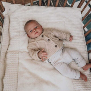 Baby lying on a white blanket in a crib, wearing a cream cardigan and light pants, looking at the camera.