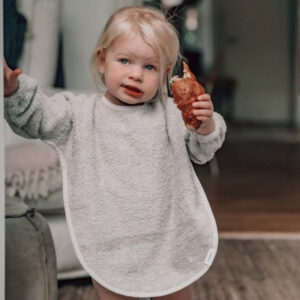 Toddler in a light gray towel bib holding a croissant and raising a hand.