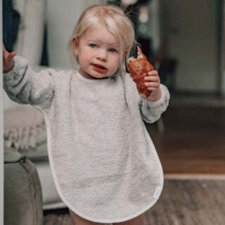 Toddler in a light gray towel bib holding a croissant and raising a hand.
