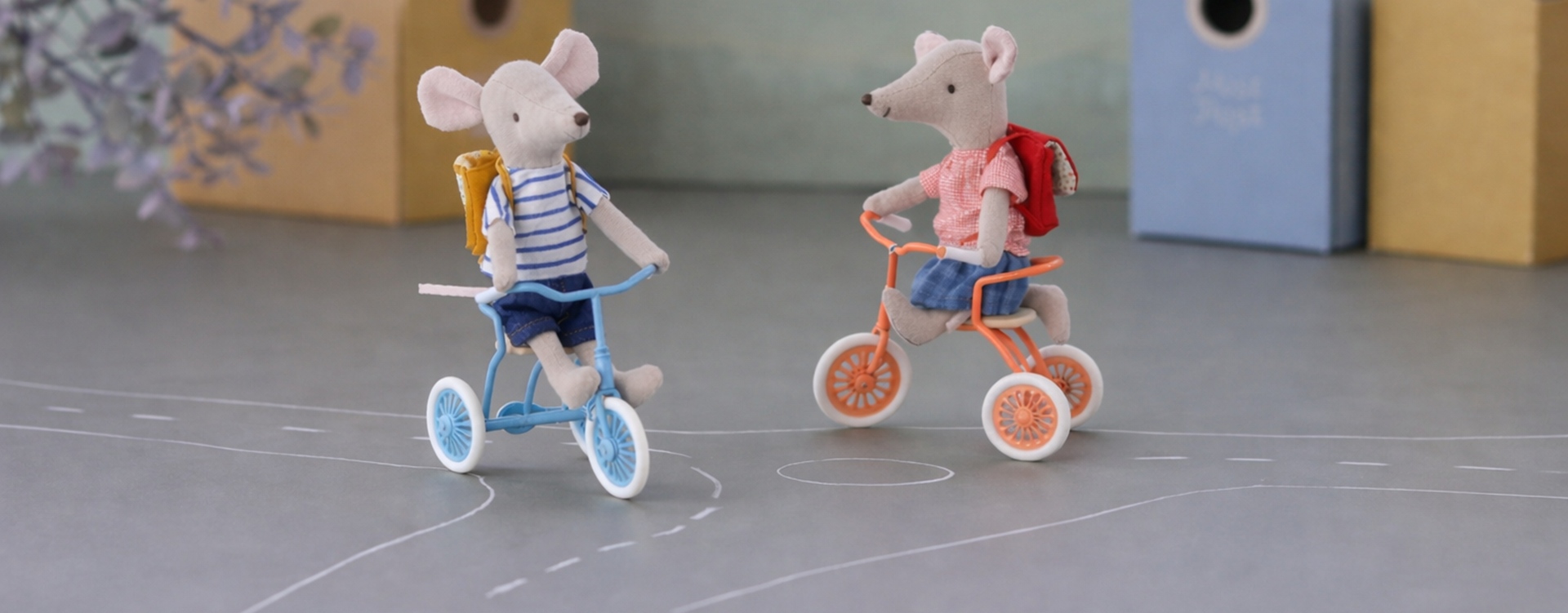 Two plush mice ride small tricycles facing each other, each with a backpack, on a chalk-drawn road indoors.