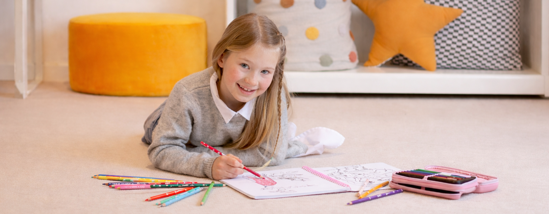 Smiling girl lying on the floor, drawing in a notebook with a red pencil while colorful pencils are scattered nearby.