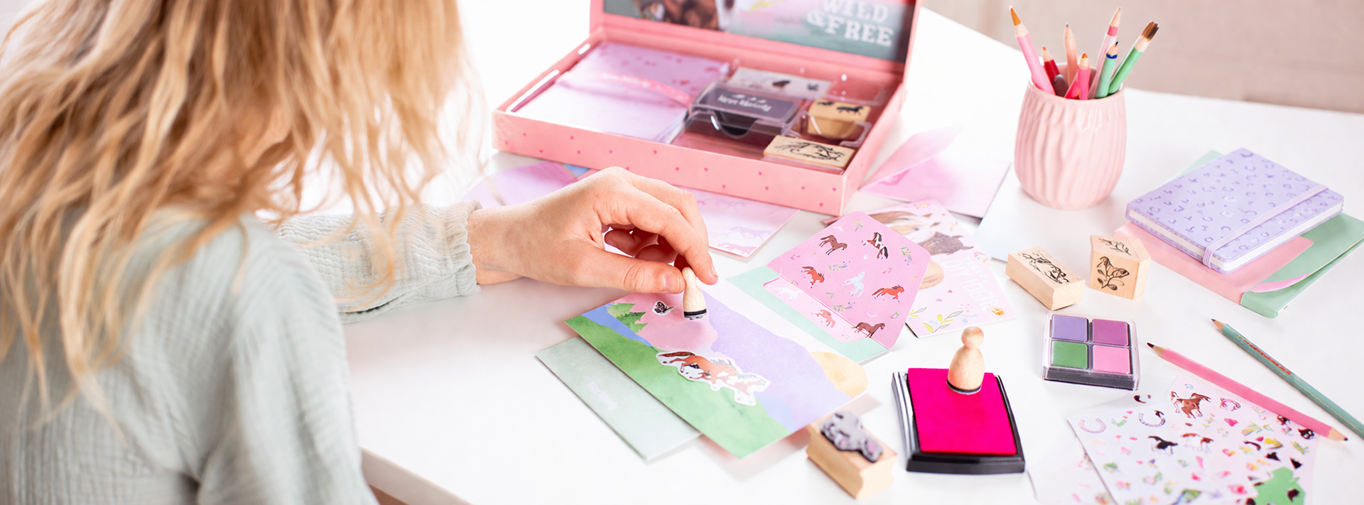 A hand stamps a pink card on a pastel craft desk, surrounded by stickers and colorful stationery.