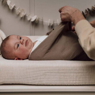 Newborn baby lying on a cushioned changing table, being swaddled in a beige blanket by an adult.