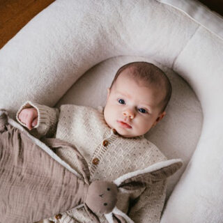 Baby lying in a soft round bassinet wearing a cream knit sweater, awake and looking up.