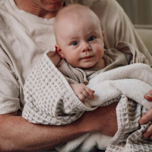 Baby wrapped in a soft blanket being held securely by an adult, looking at the camera with a gentle smile.