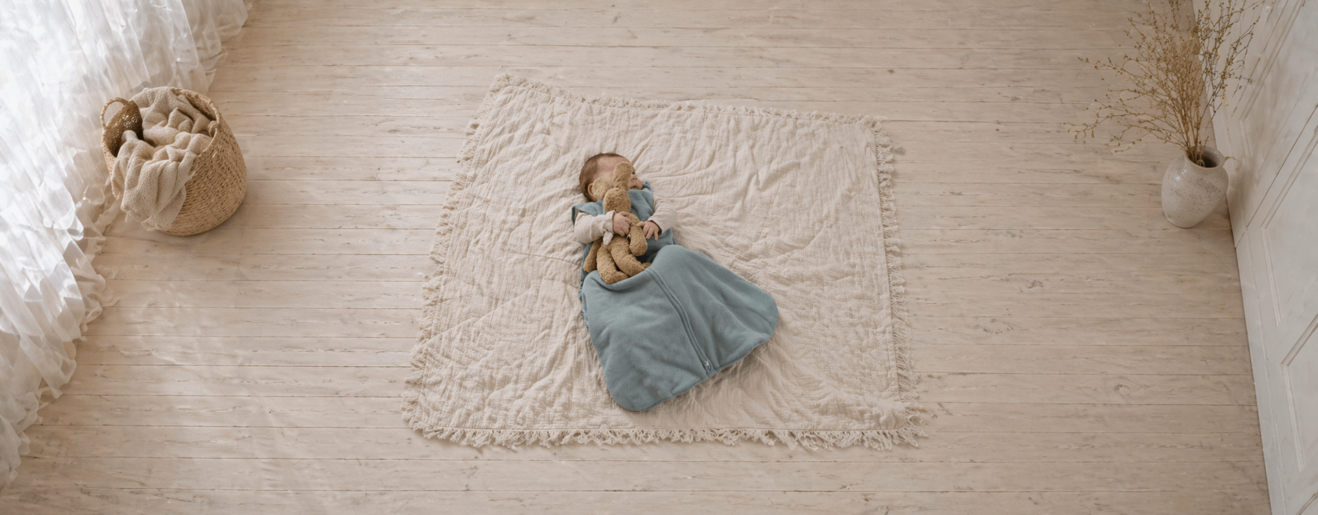 Overhead view of a baby asleep in a blue swaddle on a beige fringed rug, hugging a stuffed bear.