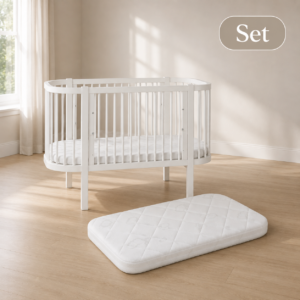 White oval baby crib with slatted sides in a bright room; a fitted mattress sits inside, and a separate foam mattress lies on the floor nearby.