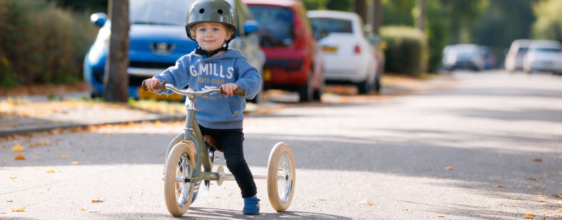 Child wearing a helmet rides a small balance bike on a sunlit autumn street with fallen leaves nearby.