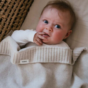 Baby lying on a light blanket in a cream knit sweater, fingers near mouth, blue eyes looking up..useful for a child-focused page