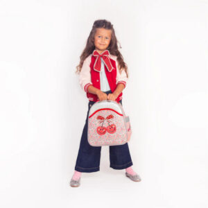 Young girl standing against a white background, holding a pastel backpack with a cherry print and red bow.