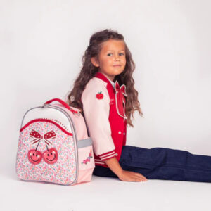 Young girl with long hair sits on the floor next to a pink cherry-patterned backpack wearing a red varsity jacket and jeans.