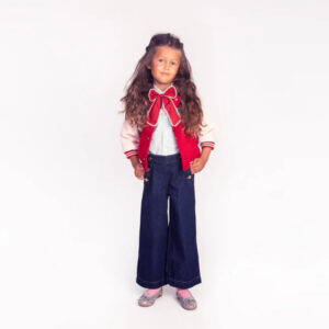 Full-body portrait of a young girl wearing a red varsity jacket, light striped shirt, wide-leg dark jeans, pink socks, and glittery shoes with a red bow at the collared neck.