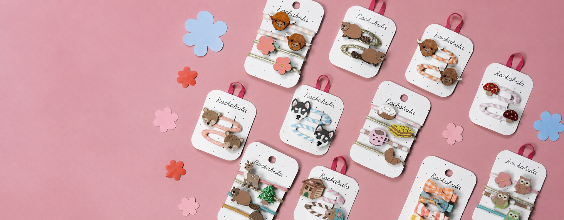 Assorted Rockahula hair accessories on white display cards arranged diagonally on a pink backdrop.