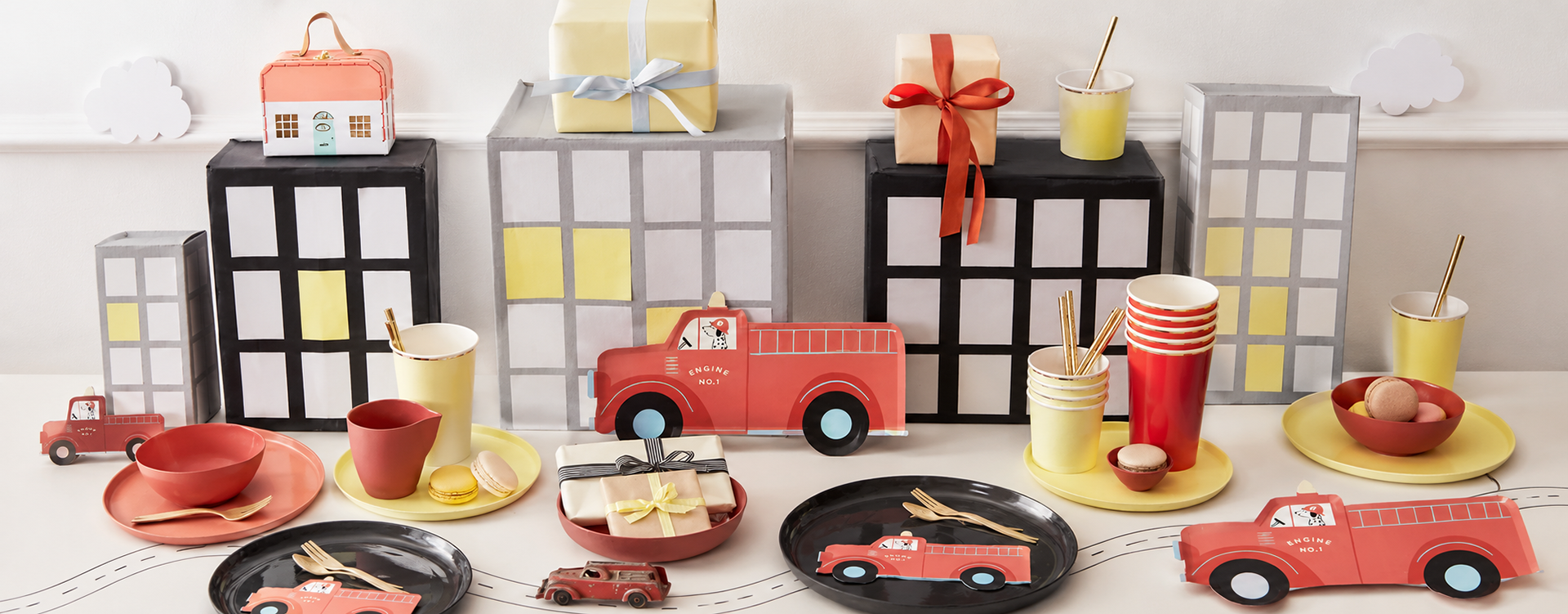 Playful party scene with red toy fire trucks, gift boxes, and colorful tableware on a white surface.
