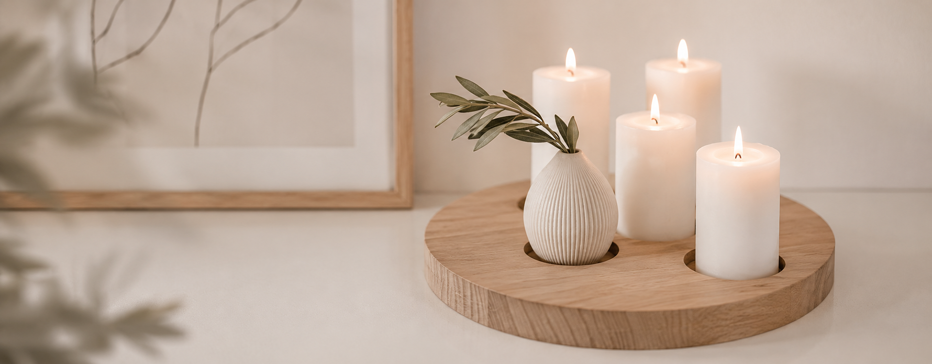 Round wooden tray holds four softly glowing white candles and a ribbed ceramic vase with green leaves, on a pale surface.