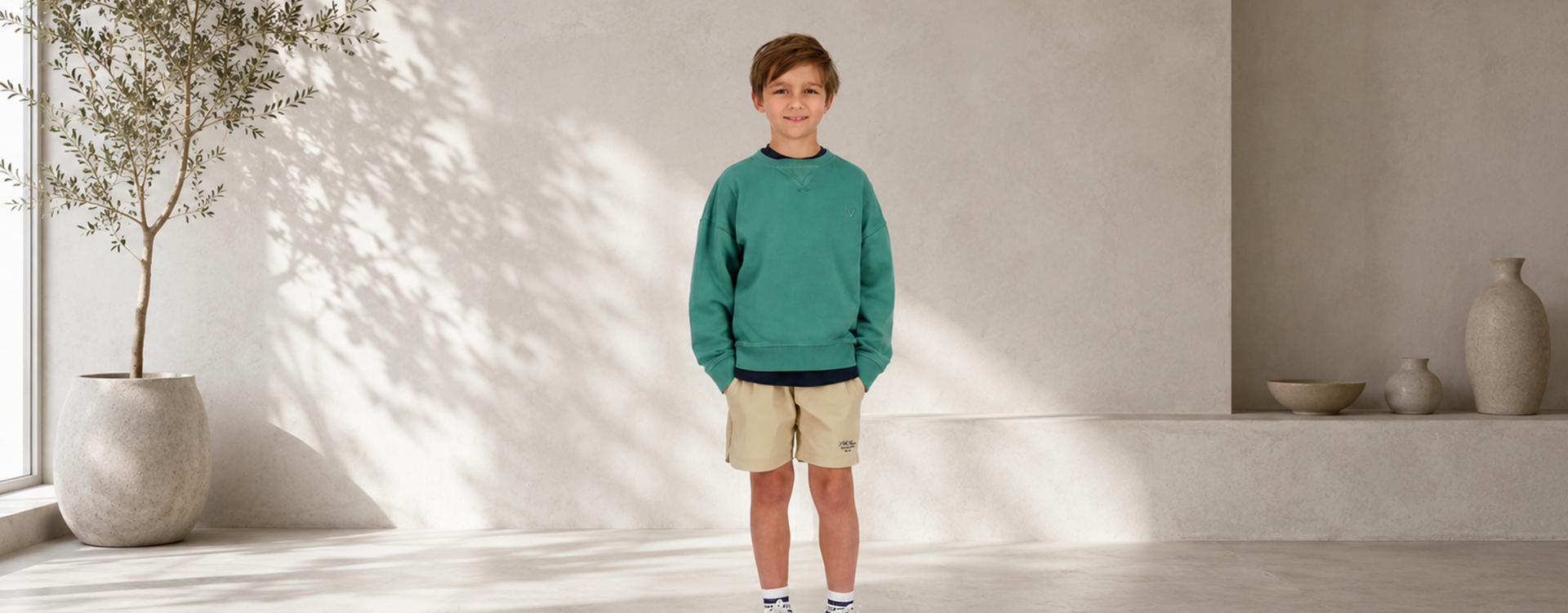 Smiling boy stands indoors in a green sweatshirt and beige shorts, hands in pockets, against a neutral beige wall.
