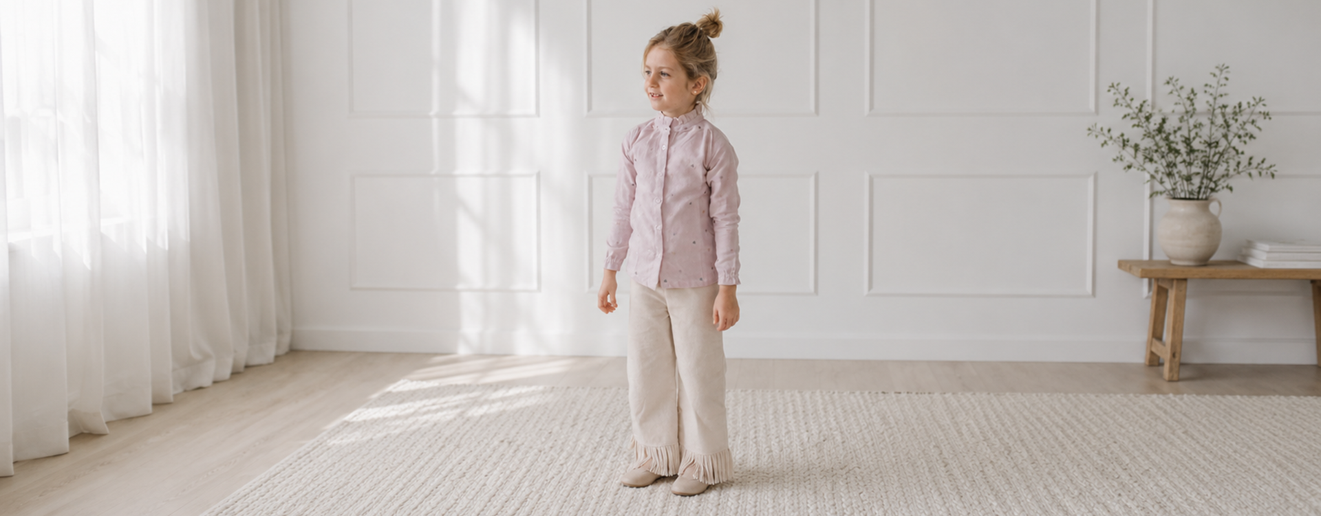 Girl standing in a bright, minimally furnished room wearing a pink shirt and beige fringed pants.