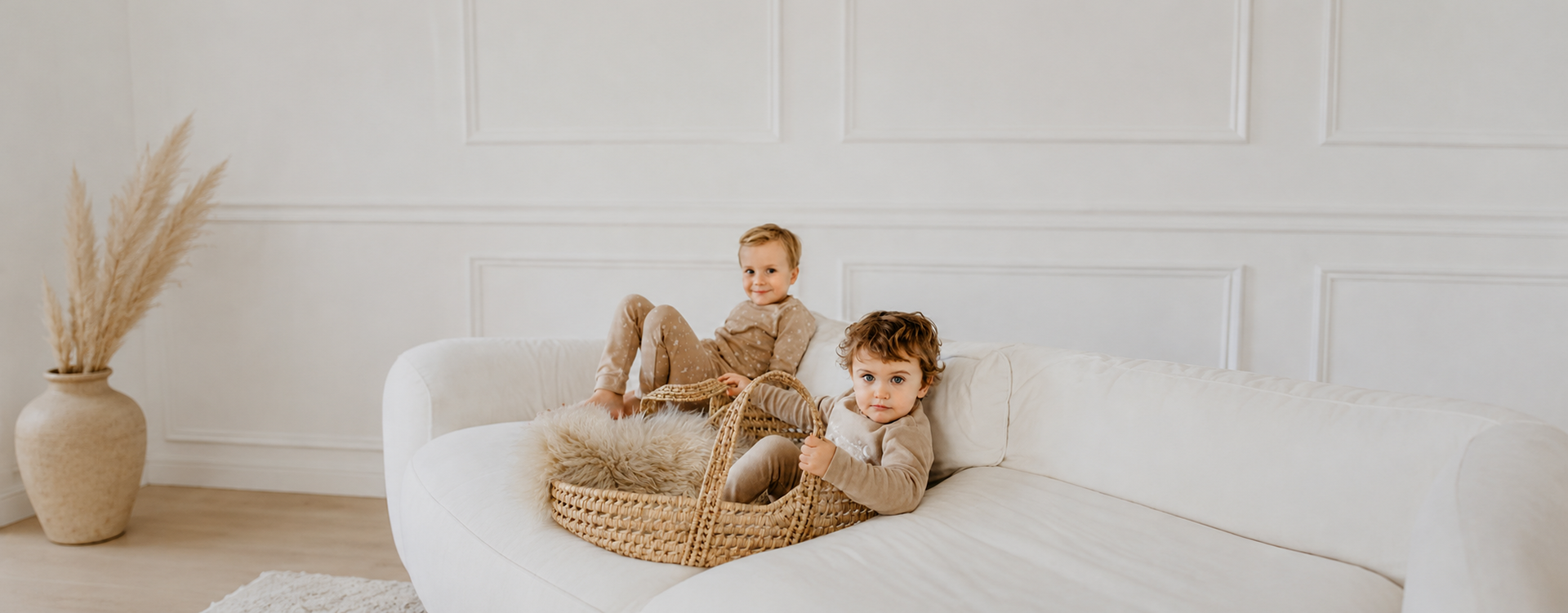 Two toddlers lounging on a wicker basket placed on a white sofa in a light, airy living room.