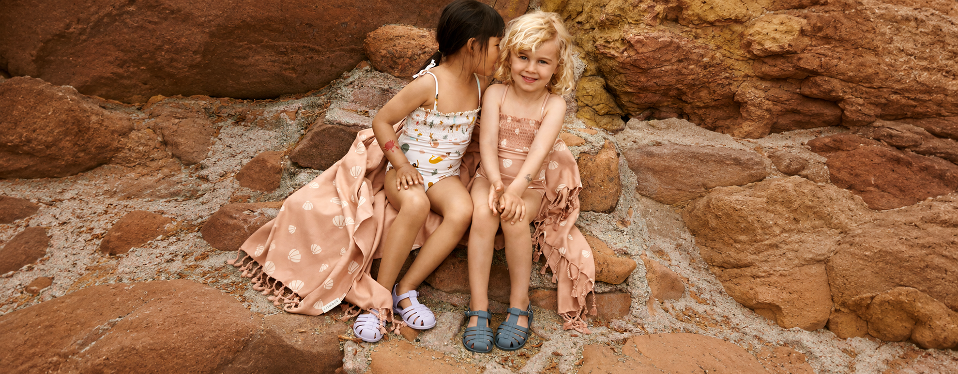 Two young girls sit close together on red rocky terrain, wearing swimsuits and sandals, smiling at the camera with a peach blanket draped beside them.