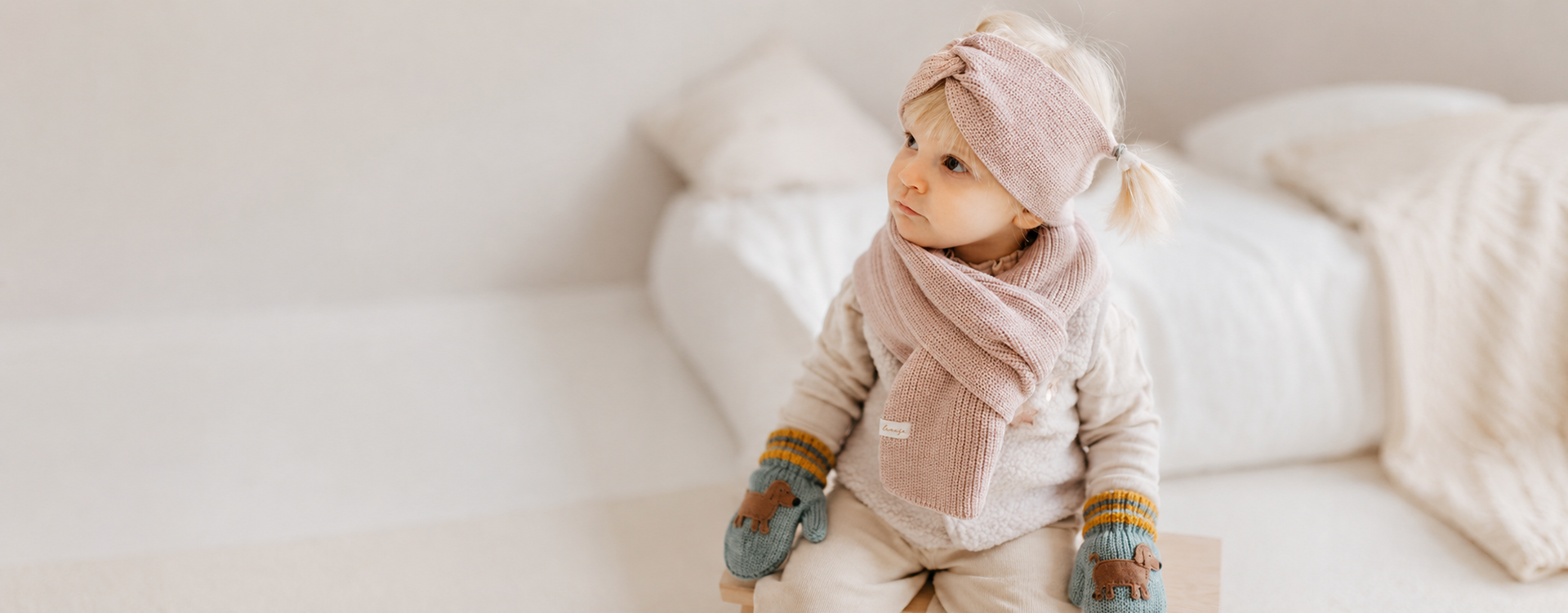 Toddler girl in pink knit headband and scarf sits indoors in a cozy beige living room, wearing teal mittens with animal patches and beige outfit.