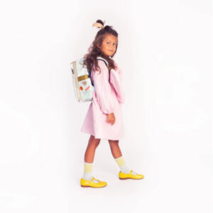 Young girl in a pink dress with yellow shoes and striped socks, wearing a pastel backpack, standing and looking toward the camera.