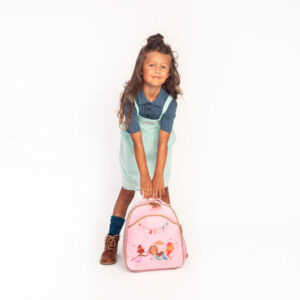 Young girl in a teal pinafore over a navy shirt, bending to hold a pink cartoon-themed backpack against a white background.