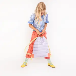 Young girl wearing a blue gingham top and red pants stands while holding a lavender drawstring backpack with yellow straps.
