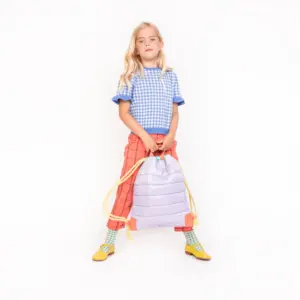 Young girl standing against a white background, holding a lavender puffed backpack with yellow drawstrings.