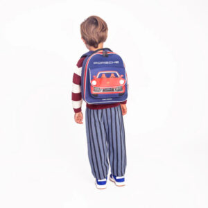 Child with back to camera wearing a blue Porsche backpack featuring a red car, striped pants, and blue sneakers.
